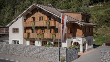 Ein charmantes Gebäude im alpinen Stil mit Holzverkleidung und Balkonen. Es steht an einer Straße, umgeben von einer Steinmauer und viel Grün.