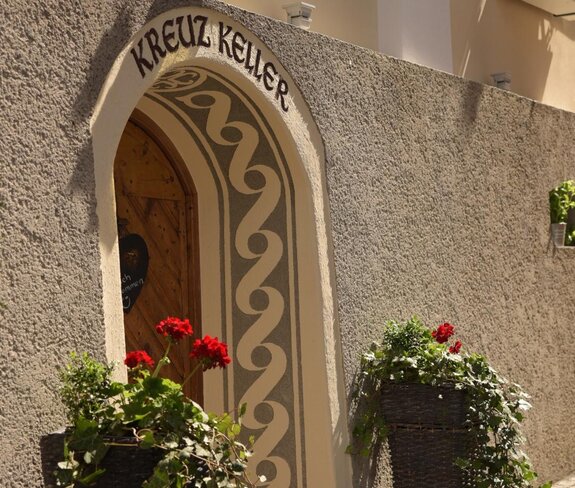A picturesque entrance with the inscription "KREUZ KELLER". Decorative flower pots with red flowers and green foliage stand in front of the entrance.