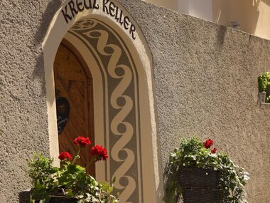 A picturesque entrance with the inscription "KREUZ KELLER". Decorative flower pots with red flowers and green foliage stand in front of the entrance.