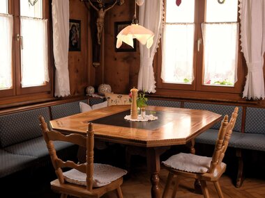 A cozy dining area with a round wooden table and chairs. The walls are made of wood, and large windows let in plenty of light.