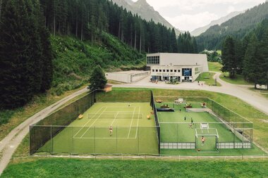 Blick auf die Sportanlage in Gargellen mit Tennisplatz und multifunktionalem Spielfeld, auf denen mehrere Personen aktiv sind. Im Hintergrund liegen Gebäude zwischen dichten Bergwäldern, darüber erheben sich die markanten Gipfel der Gargellner Bergwelt. | © Gargellner Bergbahnen GmbH & Co KG, Daniel Zangerl