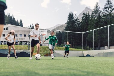 Vier Kinder spielen gemeinsam Fußball auf dem Funcourt in Gargellen. Im Hintergrund sind die umliegenden Bergwälder und Gipfel der Gargellner Bergwelt zu sehen. | © Gargellner Bergbahnen GmbH & Co KG, Daniel Zangerl