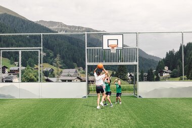 Drei Kinder spielen gemeinsam Basketball auf dem Funcourt in Gargellen. Ein Mädchen hält den Ball über dem Kopf, während zwei jüngere Kinder versuchen, ihn zu erreichen. Im Hintergrund sind der Basketballkorb, Gargellen sowie die bewaldete Berglandschaft zu sehen. | © Gargellner Bergbahnen GmbH & Co KG, Daniel Zangerl