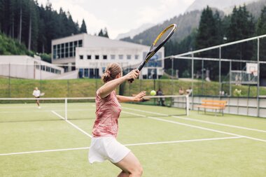 Eine Frau holt auf dem Tennisplatz in Gargellen mit ihrem Schläger zum Schlag aus, während der Tennisball auf sie zufliegt. Auf der gegenüberliegenden Platzhälfte steht eine weitere Spielerin. Im Hintergrund rahmen bewaldete Berghänge die Sportanlage ein. | © Gargellner Bergbahnen GmbH & Co KG, Daniel Zangerl
