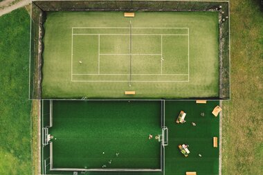 Luftaufnahme der Sportanlage in Gargellen mit einem Tennisplatz und dem angrenzenden Funcourt. Auf beiden Spielflächen sind mehrere Personen sportlich aktiv. Grünflächen und Wege umgeben die Anlage. | © Gargellner Bergbahnen GmbH & Co KG, Daniel Zangerl