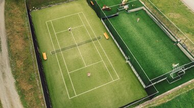 Luftaufnahme des Tennisplatzes und Funcourts in Gargellen. Zwei Personen spielen Tennis, während auf dem angrenzenden Funcourt mehrere Kinder und Erwachsene aktiv sind. Die beiden grünen Spielflächen sind von Wiesen und Wegen umgeben. | © Gargellner Bergbahnen GmbH & Co KG, Daniel Zangerl