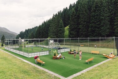 Blick auf den Funcourt und Tennisplatz in Gargellen, umgeben von grünen Wiesen und dichtem Bergwald. Kinder spielen auf dem Funcourt, während auf dem Tennisplatz gespielt wird und Besucherinnen und Besucher im angrenzenden Aufenthaltsbereich verweilen. | © Gargellner Bergbahnen GmbH & Co KG, Daniel Zangerl