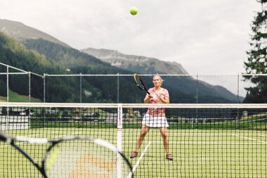 Tennisspielerin auf dem Tennisplatz in Gargellen, die sich auf den heranfliegenden Ball vorbereitet. Im Hintergrund erstrecken sich bewaldete Berghänge und die alpine Landschaft. | © Gargellner Bergbahnen GmbH & Co KG, Daniel Zangerl