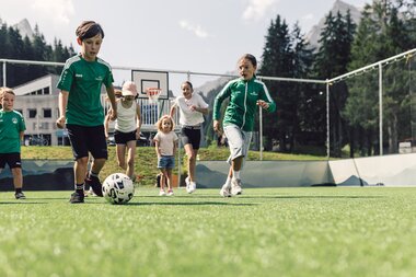 Mehrere Kinder spielen gemeinsam Fußball auf dem Funcourt in Gargellen. Im Hintergrund sind ein Basketballkorb, bewaldete Berghänge und die alpine Landschaft zu sehen. | © Gargellner Bergbahnen GmbH & Co KG, Daniel Zangerl