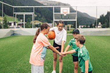 Vier Kinder stehen mit einem Basketball im Funcourt in Gargellen zusammen und legen ihre Hände übereinander. Im Hintergrund sind ein Basketballkorb sowie die Berglandschaft rund um Gargellen zu sehen. | © Gargellner Bergbahnen GmbH & Co KG, Daniel Zangerl