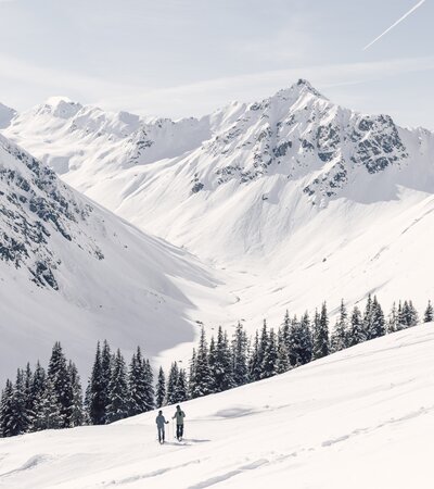 Zwei Skifaher ziehen ihre Spuren durch eine verschneite Winterlandschaft in Gargellen. Sie bewegen sich am Rand eines weiten, weißen Berghangs oberhalb eines Nadelwaldes. Im Hintergrund erstreckt sich das imposante Valzifenztal, eingerahmt von steil aufragenden, schneebedeckten Gipfeln. Der Himmel ist klar und hell, durchzogen von einem Kondensstreifen – ein ruhiger, kraftvoller Moment inmitten der Natur. | © Gargellner Bergbahnen, Daniel Zangerl