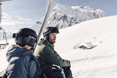 Zwei Skifahrer, eine Frau und ein Mann, sitzen gemeinsam in einem Sessellift und fahren den verschneiten Berg hinauf. Beide tragen Skibekleidung, Helme und Skibrillen. Im Hintergrund erstreckt sich eine tiefverschneite Winterlandschaft mit einem kleinen, fast eingeschneiten Holzhaus und einem imposanten, schneebedeckten Bergmassiv, der Madrisa. Die Stimmung ist ruhig und sonnig, der Himmel klar. | © Gargellner Bergbahnen GmbH & CO KG, Daniel Zangerl