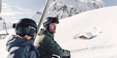 Zwei Skifahrer, eine Frau und ein Mann, sitzen gemeinsam in einem Sessellift und fahren den verschneiten Berg hinauf. Beide tragen Skibekleidung, Helme und Skibrillen. Im Hintergrund erstreckt sich eine tiefverschneite Winterlandschaft mit einem kleinen, fast eingeschneiten Holzhaus und einem imposanten, schneebedeckten Bergmassiv, der Madrisa. Die Stimmung ist ruhig und sonnig, der Himmel klar. | © Gargellner Bergbahnen GmbH & CO KG, Daniel Zangerl