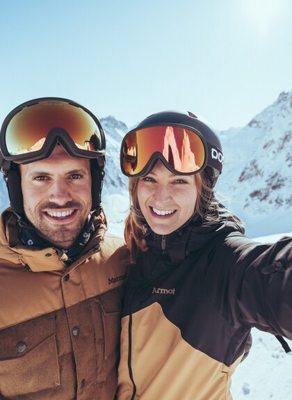 Mann und Frau im Skioutfit machen ein Selfie auf einer sonnigen Skipiste im Skigebiet Gargellen im Montafon, mit verschneiten Madrisa, so heißt der Berg im Hintergrund. | © Gargellner Bergbahnen GmbH & Co KG, Christoph Schöch 