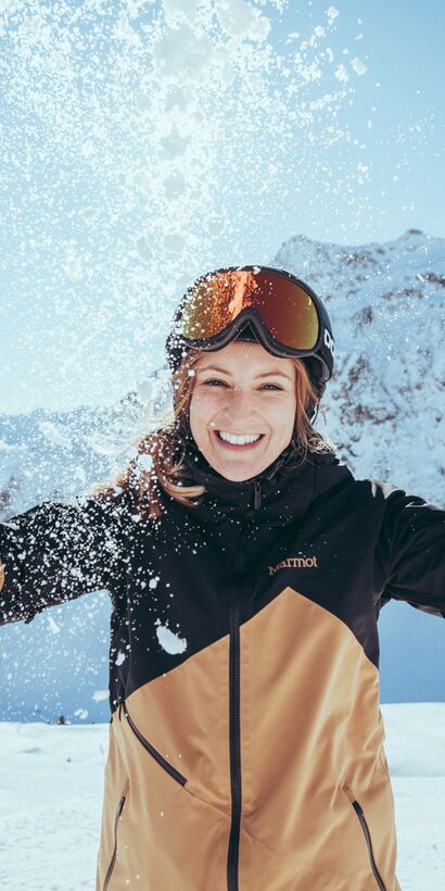 Eine lächelnde Frau in Skikleidung wirft Schnee in die Luft. Sie steht im Sonnenschein vor einer verschneiten Bergkulisse in Gargellen und genießt den Moment. | © Gargellner Bergbahnen GmbH & Co KG, Christoph Schöch