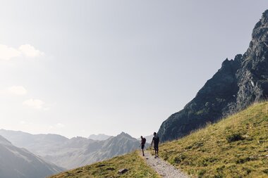 Zwei Wanderer gehen auf einem Bergpfad in Gargellen, umgeben von grünen Hängen und Felsmassiven. | © Gargellner Bergbahnen GmbH & Co KG, Daniel Zangerl