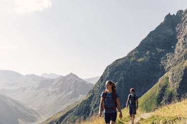 Zwei Erwachsene wandern auf einem schmalen Bergweg am Gargellner Schafberg. Im Tal und an den Hängen leuchten grüne Wiesen, umrahmt von imposanten Berggipfeln. | © Gargellner Bergbahnen GmbH & Co KG, Daniel Zangerl