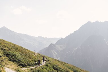 ine Familie wandert auf einem schmalen Bergweg am Gargellner Schafberg. Unter ihnen öffnet sich das grüne Vergaldental, umgeben von sanften Hängen und imposanten Gipfeln in sommerlicher Atmosphäre. | © Gargellner Bergbahnen GmbH & Co KG, Daniel Zangerl