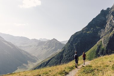 Zwei Erwachsene wandern auf einem schmalen Bergweg am Gargellner Schafberg. Im Tal und an den Hängen leuchten grüne Wiesen, umrahmt von imposanten Berggipfeln. | © Gargellner Bergbahnen GmbH & Co KG, Daniel Zangerl