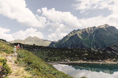 Eine Familie wandert auf einem schmalen Pfad oberhalb des Gandasees im Vergaldental. Das klare Wasser des Sees spiegelt die umliegenden Felsen und die grünen Hänge der Montafoner Berge. Die Szene zeigt eine idyllische Sommerlandschaft mit blauem Himmel und leichten Wolken. | © Gargellner Bergbahnen GmbH & Co KG, Daniel Zangerl