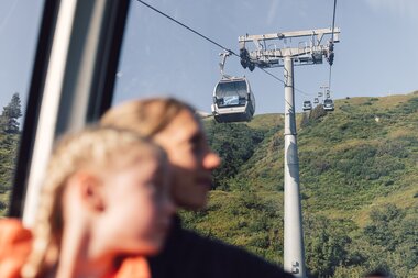 Blick aus einer Gondel auf die entgegenkommende Bergbahn über den grünen Berghängen von Gargellen. Im Vordergrund sitzen unscharf ein Kind und eine erwachsene Person in der Gondel. | © Gargellner Bergbahnen GmbH & Co KG, Daniel Zangerl