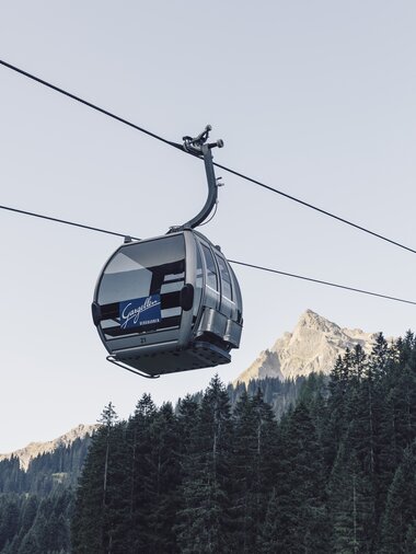 ine silberne Gondel der Gargellner Bergbahnen schwebt über einen dichten Nadelwald in Richtung Bergstation. Im Hintergrund ragt der Gipfel der Madrisa in der Abendsonne auf, während der Himmel klar und hellblau ist. | © Gargellner Bergbahnen GmbH & Co KG, Daniel Zangerl