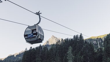 ine silberne Gondel der Gargellner Bergbahnen schwebt über einen dichten Nadelwald in Richtung Bergstation. Im Hintergrund ragt der Gipfel der Madrisa in der Abendsonne auf, während der Himmel klar und hellblau ist. | © Gargellner Bergbahnen GmbH & Co KG, Daniel Zangerl