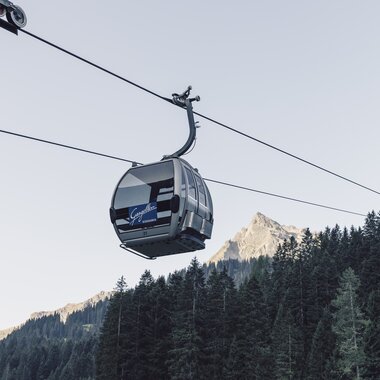 ine silberne Gondel der Gargellner Bergbahnen schwebt über einen dichten Nadelwald in Richtung Bergstation. Im Hintergrund ragt der Gipfel der Madrisa in der Abendsonne auf, während der Himmel klar und hellblau ist. | © Gargellner Bergbahnen GmbH & Co KG, Daniel Zangerl