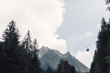 Eine Gondel der Schafbergbahn, die zu den Gargellner Bergbahnen gehört, fährt über dichte Tannenwälder in Richtung Berg. Im Hintergrund erhebt sich die imposante Madrisa unter einem teils bewölkten Sommerhimmel. | © Gargellner Bergbahnen GmbH & Co KG, Daniel Zangerl