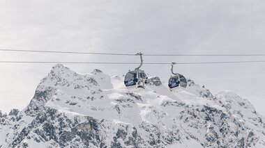 Zwei Gondeln der Schafbergbahn in Gargellen schweben vor einer beeindruckenden, schneebedeckten Bergkulisse. Die modernen Kabinen der Gargellner Bergbahnen heben sich klar vom hellen Winterhimmel ab, während im Hintergrund die majestätischen Gipfel der Montafoner Alpen aufragen. | © Gargellner Bergbahnen GmbH & Co KG, Daniel Zangerl