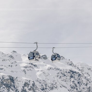 Zwei Gondeln der Schafbergbahn in Gargellen schweben vor einer beeindruckenden, schneebedeckten Bergkulisse. Die modernen Kabinen der Gargellner Bergbahnen heben sich klar vom hellen Winterhimmel ab, während im Hintergrund die majestätischen Gipfel der Montafoner Alpen aufragen. | © Gargellner Bergbahnen GmbH & Co KG, Daniel Zangerl