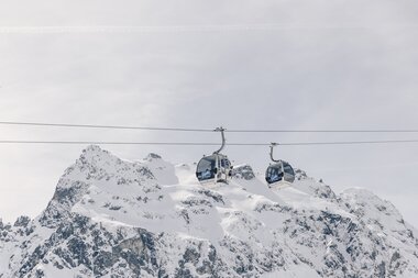 Zwei Gondeln der Schafbergbahn in Gargellen schweben vor einer beeindruckenden, schneebedeckten Bergkulisse. Die modernen Kabinen der Gargellner Bergbahnen heben sich klar vom hellen Winterhimmel ab, während im Hintergrund die majestätischen Gipfel der Montafoner Alpen aufragen. | © Gargellner Bergbahnen GmbH & Co KG, Daniel Zangerl