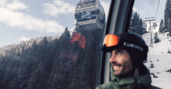 Ein Skifahrer mit Helm und Skibrille sitzt in einer Gondel der Schafbergbahn in Gargellen. Durch das Fenster ist die Seilbahn zu sehen, die über verschneite Wälder und Hänge führt. Im Hintergrund steigen die Gondeln der Gargellner Bergbahnen den schneebedeckten Berg hinauf. | © Gargellner Bergbahnen GmbH & Co KG, Daniel Zangerl
