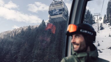 Ein Skifahrer mit Helm und Skibrille sitzt in einer Gondel der Schafbergbahn in Gargellen. Durch das Fenster ist die Seilbahn zu sehen, die über verschneite Wälder und Hänge führt. Im Hintergrund steigen die Gondeln der Gargellner Bergbahnen den schneebedeckten Berg hinauf. | © Gargellner Bergbahnen GmbH & Co KG, Daniel Zangerl