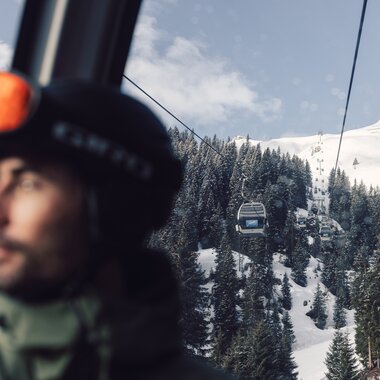 Ein Skifahrer mit Helm und Skibrille sitzt in einer Gondel der Schafbergbahn in Gargellen. Durch das Fenster ist die Seilbahn zu sehen, die über verschneite Wälder und Hänge führt. Im Hintergrund steigen die Gondeln der Gargellner Bergbahnen den schneebedeckten Berg hinauf. | © Gargellner Bergbahnen GmbH & Co KG, Daniel Zangerl