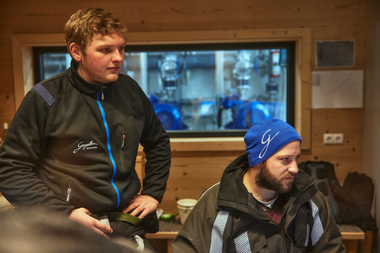Zwei Mitarbeiter der Gargellner Bergbahnen sitzen in einem holzverkleideten Technikraum und überwachen die Anlagensteuerung. Einer trägt eine blaue Mütze mit dem Schriftzug „G“, der andere eine schwarze Arbeitsjacke mit dem Logo der Gargellner Bergbahnen. Durch das Fenster im Hintergrund sind technische Geräte und Pumpen zu sehen. | © Gargellner Bergbahnen GmbH & Co KG, Stefan Kothner