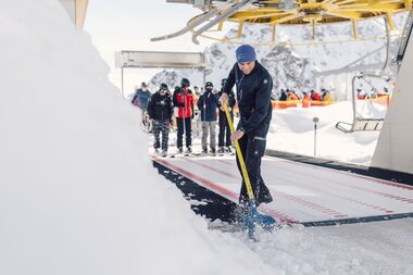 Ein Mitarbeiter der Gargellner Bergbahnen räumt mit einer Schneeschaufel den Bereich bei der Bergstation frei. Im Hintergrund warten Skifahrer auf den Einstieg in den Sessellift, umgeben von verschneiten Bergen und blauem Himmel. | © Gargellner Bergbahnen GmbH & Co KG, Daniel Zangerl