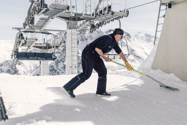 Ein Mitarbeiter der Gargellner Bergbahnen räumt Schnee an der Bergstation einer Sesselbahn. Im Hintergrund sind die verschneiten Gipfel der Gargellner Bergwelt zu sehen. | © Gargellner Bergbahnen GmbH & Co KG, Daniel Zangerl