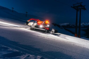 Eine beleuchtete Pistenraupe präpariert bei Nacht die Skipisten in Gargellen. Die Maschine fährt über die frisch verschneite Abfahrt, während im Hintergrund die Berggipfel und eine Seilbahnstütze zu sehen sind. Das Licht der Pistenraupe leuchtet den Schnee hell aus und schafft eine stimmungsvolle Atmosphäre. | © Montafon Tourismus, Marie Schilcher