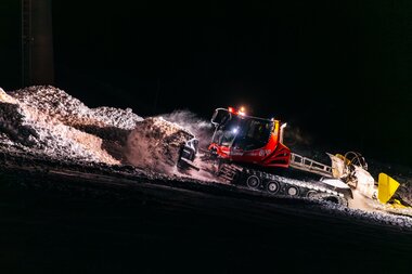 Eine Pistenraupe der Gargellner Bergbahnen arbeitet bei Nacht auf der beleuchteten Skipiste. Die Maschine schiebt und verteilt Schnee unter kräftigem Lichteinsatz, während der Schnee in der Dunkelheit glitzert. Im Vordergrund ist der aufgewirbelte Schnee sichtbar, im Hintergrund verschwindet die Piste in der Dunkelheit. | © Montafon Tourismus, Marie Schilcher