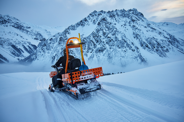 Ein Mitarbeiter der Gargellner Bergbahnen fährt mit einem orangefarbenen Motorschlitten über eine frisch verschneite Piste. Im Hintergrund ragen die verschneiten Madrisa-Berge in den Himmel, während Nebel durch das Tal zieht. | © Gargellner Bergbahnen GmbH & Co KG, Stefan Kothner