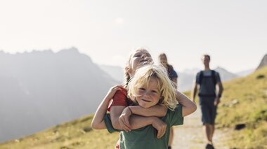Zwei Kinder umarmen sich lachend auf einem sonnigen Wanderweg in Gargellen. Im Hintergrund folgen Erwachsene, während die umliegenden Berge in sanftem Licht erstrahlen. | © Gargellner Bergbahnen, Daniel Zangerl