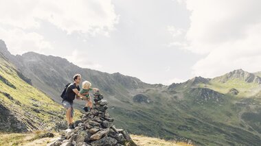 Ein Vater hilft seinem kleinen Sohn, auf ein großes Steinmandl zu steigen. Im Hintergrund erstreckt sich eine beeindruckende Berglandschaft in Gargellen, die von Sonnenlicht überflutet ist. | © Gargellner Bergbahnen, Daniel Zangerl