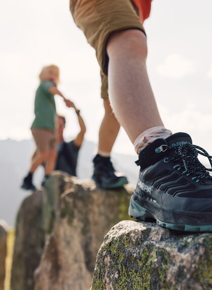 Eine Familie wandert entlang großer Felsen und balanciert spielerisch über die Steine. Die Kinder werden von den Eltern an der Hand gehalten, während im Hintergrund die Berge des Montafon leuchten. | © Gargellner Bergbahnen GmbH & Co KG, Daniel Zangerl