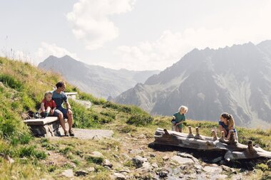 ine Familie genießt den sonnigen Tag am Wasserspielplatz in den Bergen von Gargellen. Während ein Kind mit einer Gießkanne spielt, sitzen Vater und Tochter entspannt auf einer Bank und genießen die Aussicht auf die imposante Bergkulisse. | © Gargellner Bergbahnen GmbH & Co KG, Daniel Zangerl