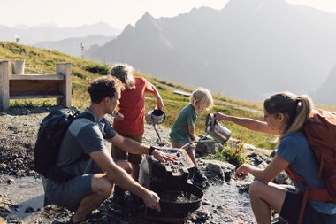 Eine Familie mit zwei Kindern wäscht gemeinsam Gold in den Bergen von Gargellen. Die Kinder gießen Wasser mit Kannen, während die Eltern beim Sieben helfen. Im Hintergrund sind grüne Wiesen und Berggipfel zu sehen. | © Gargellner Bergbahnen GmbH & Co KG, Daniel Zangerl