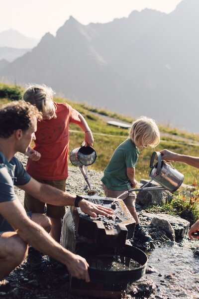 Eine Familie mit zwei Kindern wäscht gemeinsam Gold in den Bergen von Gargellen. Die Kinder gießen Wasser mit Kannen, während die Eltern beim Sieben helfen. Im Hintergrund sind grüne Wiesen und Berggipfel zu sehen. | © Gargellner Bergbahnen GmbH & Co KG, Daniel Zangerl