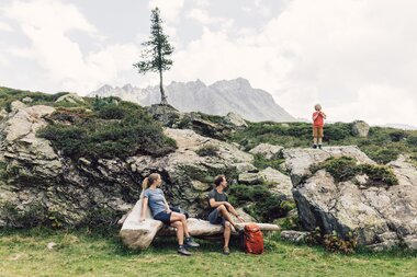 Eine Familie macht eine Pause zwischen Felsen in den Bergen bei Gargellen. Zwei Erwachsene sitzen entspannt auf einer Holzbank, während ein Kind auf einem Felsen steht und ruft. Im Hintergrund ragen die markanten Gipfel der Montafoner Alpen auf. | © Gargellner Bergbahnen GmbH & Co KG, Daniel Zangerl