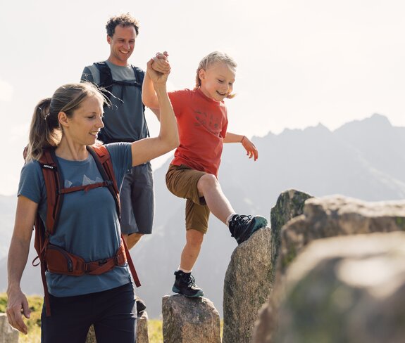 Eine Familie überquert große Felsen in den Bergen von Gargellen. Die Mutter hält die Hand ihres Kindes, während der Vater lächelnd folgt – ein Moment voller Vertrauen und Freude beim Wandern in der Sonne. | © Gargellner Bergbahnen GmbH & Co KG, Daniel Zangerl