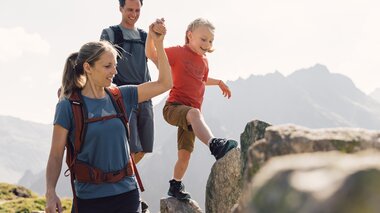 Eine Familie überquert große Felsen in den Bergen von Gargellen. Die Mutter hält die Hand ihres Kindes, während der Vater lächelnd folgt – ein Moment voller Vertrauen und Freude beim Wandern in der Sonne. | © Gargellner Bergbahnen GmbH & Co KG, Daniel Zangerl